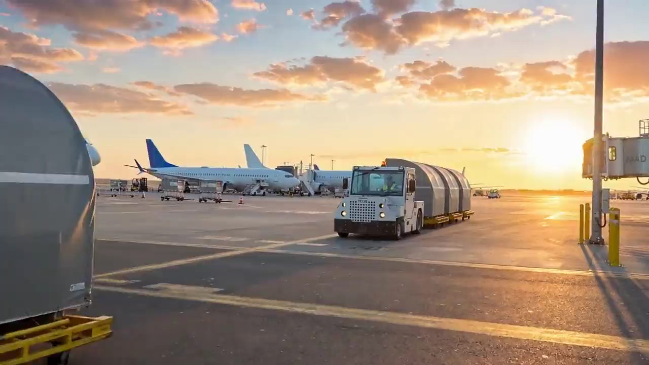 A tug and driver pulling a train of ULD containers across the ramp at sunset, an aircraft behind